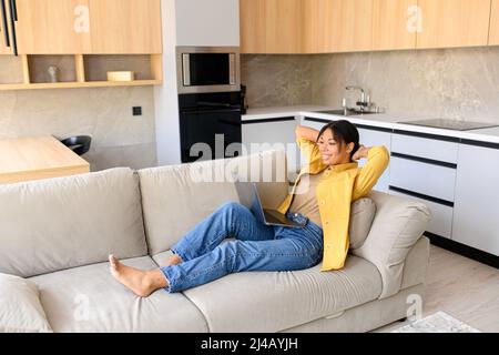 Sereine femme afro-américaine utilisant un ordinateur portable pour travailler ou étudier à distance, assise sur le canapé et se reposer, tenant les mains derrière la tête, passe un week-end paresseux à la maison, rêvant de jour Banque D'Images