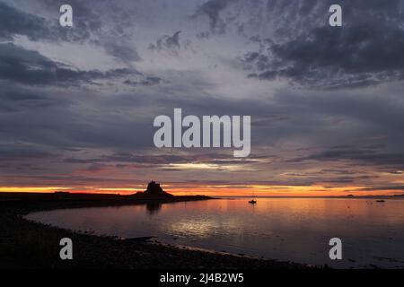 Voile et bateau à moteur dans le port de l'île Sainte. Des moments de marée haute avant le lever du soleil dans un ciel rouge, jaune et orange vif. Château de Bamburgh à l'horizon Banque D'Images