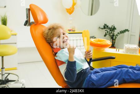 Un garçon mignon est assis dans une chaise dentaire dans une clinique moderne. Un bébé charmant regarde dans un miroir d'endurance. Le concept de la dentisterie pédiatrique Banque D'Images