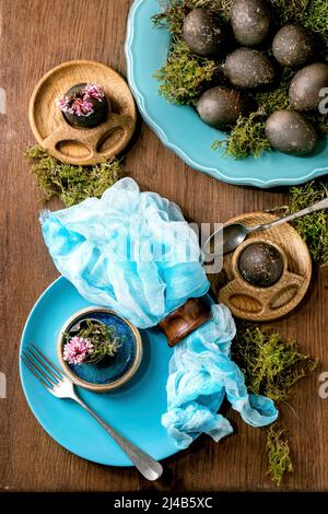 Table de Pâques avec assiettes en céramique turquoise et coquetiers vides, décorée de mousse forestière, œufs de pâques de couleur noire et fleurs de printemps roses Banque D'Images