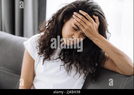 La jeune femme fatiguée est assise sur le canapé, ayant un mal de tête. La femme afro-américaine malheureuse est malsaine, stressée, bouleversée par la maladie ou a des problèmes Banque D'Images