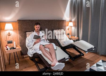 Un jeune homme charmant lisant le magazine tout en se reposant sur une chaise longue dans la chambre d'hôtel Banque D'Images