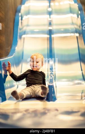 Un petit enfant qui fait du toboggan pour enfants dans la salle de jeux. Joyeux petit enfant, enfants monter, descendre sur le toboggan, dans le centre de jeux, amusant. Banque D'Images