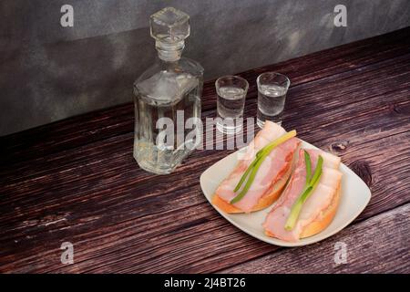 Une carafe en verre et deux verres de vodka russe et une assiette avec deux sandwiches à la viande fumée sur une table en bois. Gros plan. Banque D'Images