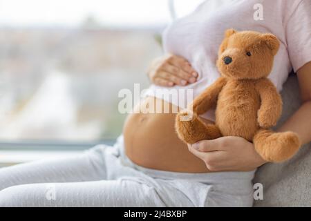 S'attendre à un bébé. Femme enceinte embrassant l'estomac et l'ours en peluche jouet heureux attendant la naissance d'un enfant. Planification familiale. Grossesse naturelle à la maison Banque D'Images