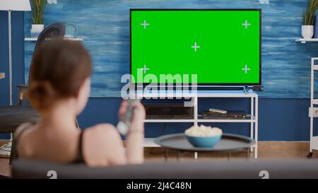 Trépied statique d'une jeune femme qui bascule entre les chaînes tout en regardant l'écran vert à la télévision et assis sur un canapé. Vue sur l'épaule d'une fille se relaxant à l'aide de la télécommande de télévision zapping sur l'affichage de clé chroma. Banque D'Images