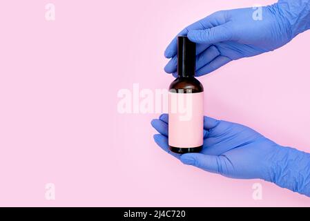Flacon cosmétique pour l'huile ou un agent antibactérien avec un endroit pour le texte dans les mains dans des gants stériles sur fond rose. Coronavirus, désinfectants Banque D'Images