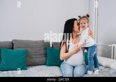 Mère et fille gaies sont sur le canapé. Le visage d'une maman enceinte s'est tourné vers le visage de sa fille et une petite fille avec des queues de cheval regarde l'appareil photo. Banque D'Images