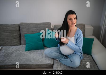 Elle est confortablement installée dans la salle de séjour, elle s'assoit sur le canapé et se peigne ses longs cheveux noirs pendant qu'elle regarde directement l'appareil photo. Banque D'Images