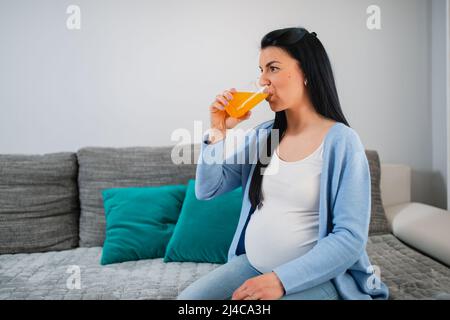 Une femme blanche enceinte en bonne santé boit du jus d'orange frais mélangé et prend les vitamines nécessaires. Une femme aux cheveux noirs est assise sur un canapé dans le salon. Banque D'Images