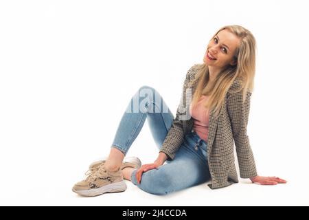 Souriant gai belle jeune femme caucasienne regardant l'appareil photo tout en s'asseyant dans un studio sur fond blanc. Sport mode décontracté. Photo de haute qualité Banque D'Images