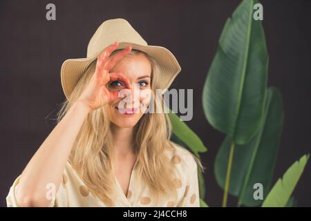 Drôle de caucasien blonde jeune fille debout devant une plante de grosse feuille dans un chapeau faisant un geste ok avec la main souriante, oeil regardant à travers les doigts avec le visage heureux. Concept de voyageur. Safari. Photo de haute qualité Banque D'Images