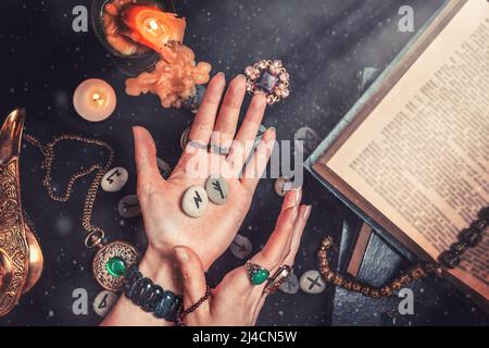 Astrologie et ésotéricisme. La sorcière tient dans ses mains les pierres de la rune.mains de près. Sur un fond noir se trouvent des runes qui disent de la fortune, un livre, Banque D'Images