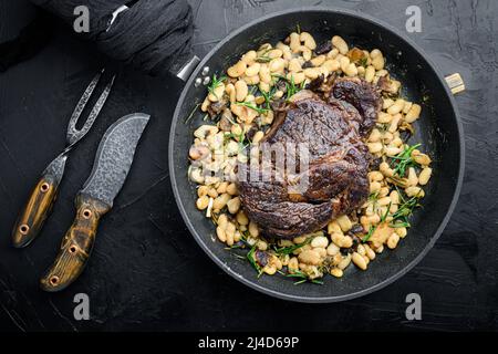 Steak d'oeil de côte avec des herbes, haricots rôtis, thym et ail, sur poêle en fonte à frire, avec couteau à viande et fourchette, sur fond de pierre noire, vue de dessus à plat Banque D'Images