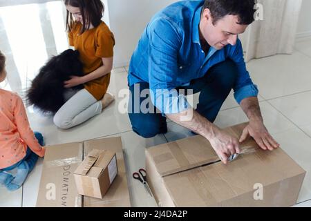 Les enfants aident leur père à emballer des boîtes en carton pour le déplacement. Jeune homme concentré mettant du ruban adhésif sur une boîte en carton pendant que toute la famille prépare t Banque D'Images