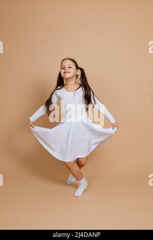 Jeune fille souriante heureuse avec de longs cheveux foncés en robe blanche, chaussettes et chaussures de gymnastique debout sur les orteils, faire curtsy, tenant la robe avec les mains sur Banque D'Images