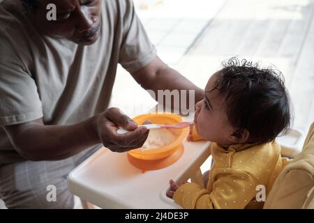 Vue en grand angle d'un homme afro-américain mûr assis devant sa petite fille qui la nourrit de purée Banque D'Images