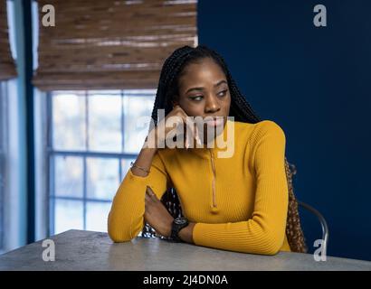 Young African American Woman avec chemise jaune assis au comptoir de la cuisine de l'île. Image style ethnique très tendance. Banque D'Images