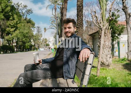 Posant, jeune homme posant pour l'appareil photo profitant du beau temps, il tient une tasse de café avec un ciel bleu profond dans l'arrière-plan Banque D'Images