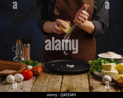 Un chef dans un uniforme sombre râpe du fromage sur une râpe à main sur une table de cuisine en bois. Légumes, oeufs, herbes, épices sur la table, fond bleu foncé. Banque D'Images