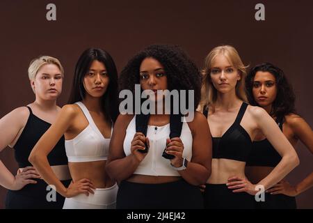 Différentes femmes dans les vêtements de sport regardant la caméra tout en se tenant sur un fond marron. Cinq femelles de différentes courses en studio. Banque D'Images