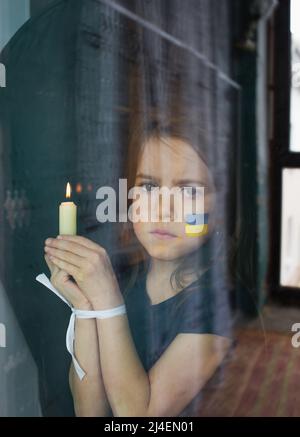 Une triste petite fille ukrainienne avec un drapeau peint sur son visage est debout derrière le verre avec ses mains attachées et tenant une bougie dans sa main. Le chi Banque D'Images