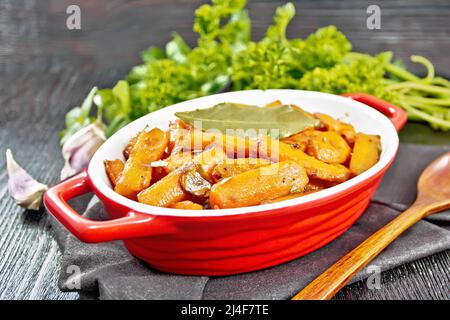 Carottes frites à l'ail, feuilles de Laurier, épices et oignons verts dans une casserole sur une serviette, basilic et persil sur fond de bois foncé Banque D'Images