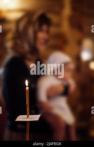 Longue bougie avec feuille de papier brûlant près d'une femme portant un foulard, tenant bébé dans des vêtements blancs dans l'église orthodoxe. Banque D'Images