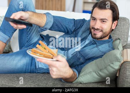 homme sur le canapé tenant le sandwich et la télécommande Banque D'Images