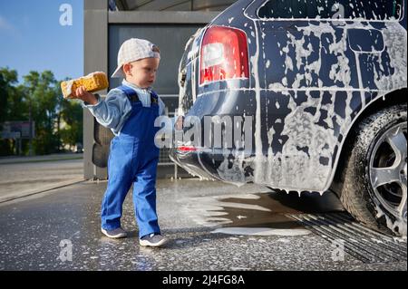 Enfant garçon étudiant le travail au lavage de voiture. Enfant en bas âge, tenant une éponge en mousse jaune dans sa main droite, examinant le wagon-savon et se préparant à commencer à nettoyer la partie arrière du transport. Banque D'Images