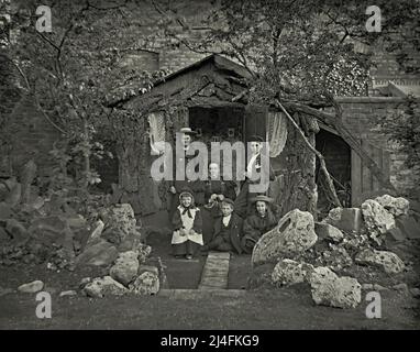 Un groupe familial avec une mère et cinq enfants pose devant leur maison d'été victorienne en Grande-Bretagne vers 1900. La maison d'été rustique dispose de fenêtres avec des rideaux en filet, du papier peint et des images sur le mur. À côté d'elle est un hangar avec l'espace de stockage. Devant la famille se trouve un impressionnant jardin de rochers avec des rochers et des rochers massifs. Ceci est tiré d'un négatif en verre victorien – une photographie vintage 1800s/1900s. Banque D'Images