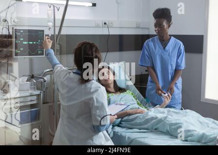 Le médecin met en place un dispositif de rattrapage de ligne centrale avec un médicament IV goutte à goutte pour le patient pendant que l'infirmière met l'oxymètre de pouls sur le doigt. Femme dans le lit d'hôpital regardant le médecin donnant ses antibiotiques intraveineux. Banque D'Images