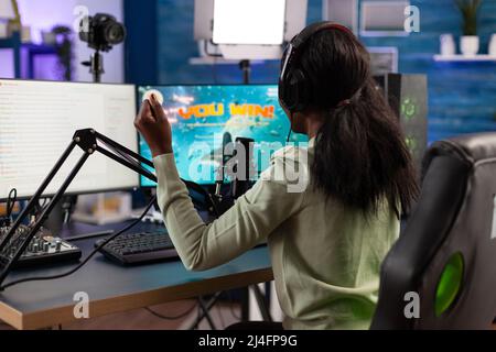 Bonne femme joueuse réussie portant des écouteurs gagnant jeux vidéo compétition parlant dans le microphone avec les joueurs. Un joueur enthousiaste jouant à des jeux de tir spatial en utilisant un ordinateur RVB. Diffusion en direct Banque D'Images