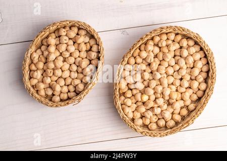 Pois chiches bio non cuits dans deux bols en paille, gros plan, sur une table en bois blanc, vue du dessus. Banque D'Images