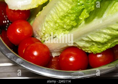 Gros plan de petites tomates fraîches et de feuilles de laitue lavées dans une passoire en métal. Tomates sur le fond en mise au point sélective. Banque D'Images