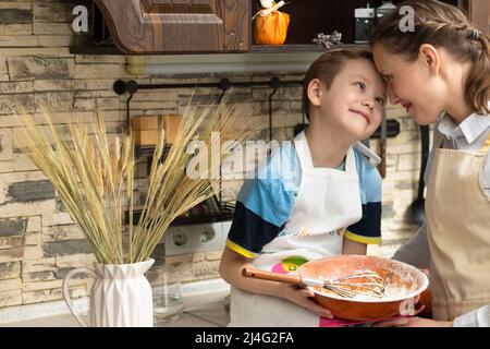 Belle jeune maman cuisinier dans un tablier avec son fils prépare la pâte à biscuits dans un bol en bois à la maison dans la cuisine. Mise au point sélective. Portrait. Gros plan Banque D'Images