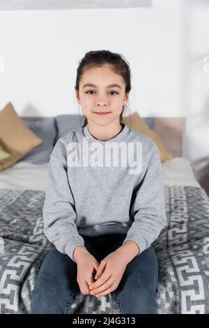préteen souriant à l'appareil photo tout en étant assise sur un lit flou Banque D'Images