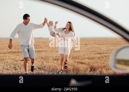 Vue à travers la fenêtre de voiture, maman et papa tenant leur fille très jeune par les mains tout en marchant sur le terrain au coucher du soleil Banque D'Images