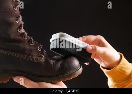 La femme nettoie et applique la crème sur les bottes d'hiver nubuck brunes pour femmes avec une éponge. Sur fond noir, gros plan Banque D'Images