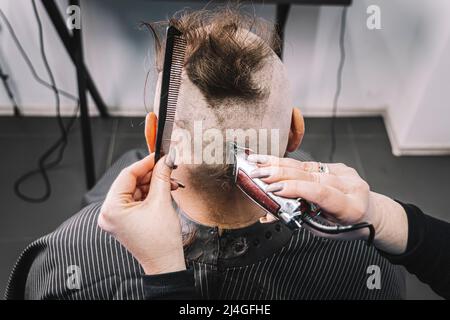 Jeune homme barbu avec un mohawk se couper la cheveux par un coiffeur tout en étant assis dans une chaise au salon de coiffure. Une fille de coiffeur fait un mohawk à un type. Banque D'Images