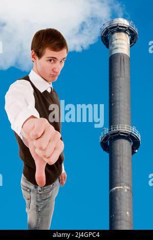 Taille haute vue d'un jeune homme blanc montrant les pouces vers le bas à côté d'une cheminée d'usine de fumeurs contre un ciel bleu. Banque D'Images