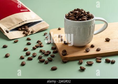 Grains de café torréfiés dans une petite tasse blanche sur fond vert pastel. Grains de café entiers déversés d'un sac de papier brun. Grains de café biologiques. Banque D'Images