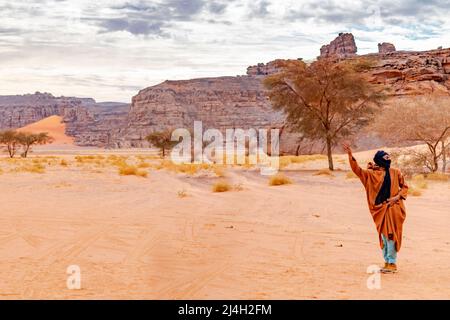 Tadraart Rouge, tuareg homme lève le bras. Acacia, sable dégradé de couleur, herbes sèches, pierres de sable jaune, montagne rocheuse et ciel couvert gris nuageux Banque D'Images