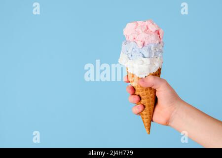 Crème glacée dans un cône avec trois saveurs différentes tenant la main d'un enfant sur un fond bleu. Concept de crème glacée avantages pour la santé Banque D'Images