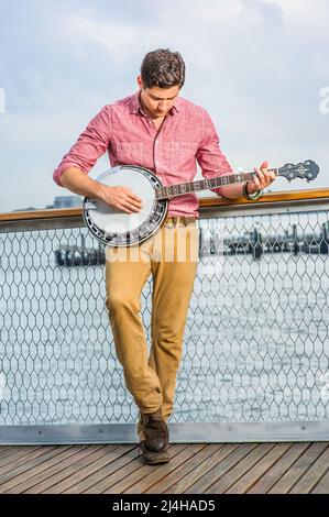 Avec sa chemise rouge clair, son pantalon jaune foncé et ses chaussures brunes, un jeune musicien se pose sur le banjo Banque D'Images