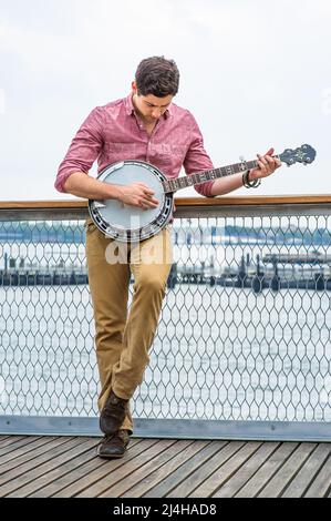 Avec sa chemise rouge clair, son pantalon jaune foncé et ses chaussures brunes, un jeune musicien se pose sur le banjo Banque D'Images