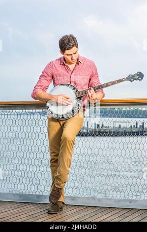 Avec sa chemise rouge clair, son pantalon jaune foncé et ses chaussures brunes, un jeune musicien se pose sur le banjo Banque D'Images