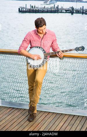Avec sa chemise rouge clair, son pantalon jaune foncé et ses chaussures brunes, un jeune musicien se pose sur le banjo Banque D'Images