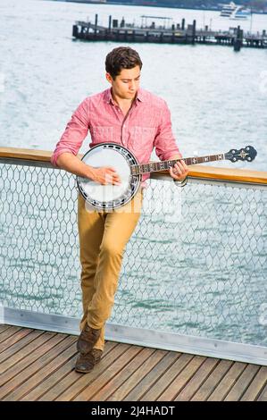 Avec sa chemise rouge clair, son pantalon jaune foncé et ses chaussures brunes, un jeune musicien joue un banjo sur le pont Banque D'Images