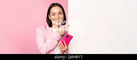 Image d'une femme d'entreprise asiatique souriante en costume, en tenant un smartphone, en pointant vers le tableau, en montrant un graphique ou un logo d'information sur un mur blanc vide, debout Banque D'Images